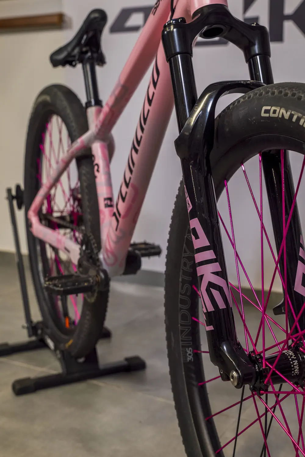 Różowy rower górski z folią ochronną na ramie, ustawiony na stojaku, z akcentami w kolorze fuksji